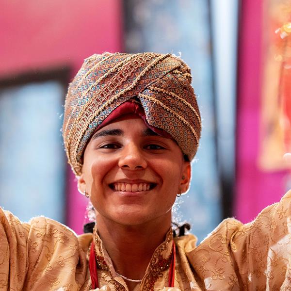 Teenager in a turban smiling into the camera between two folds of bright coloured and sequined cloth.