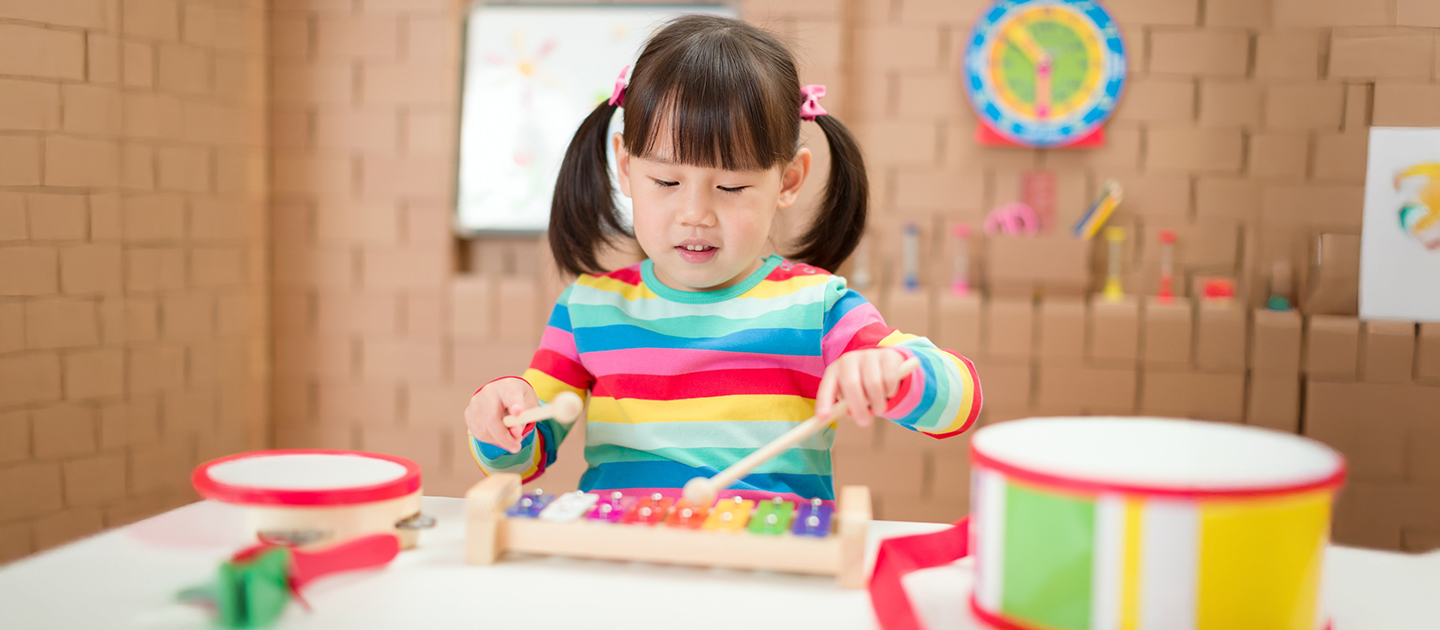 Young girl in a bright striped jumper playing a coloured xylophone, surrounded by musical instruments. 