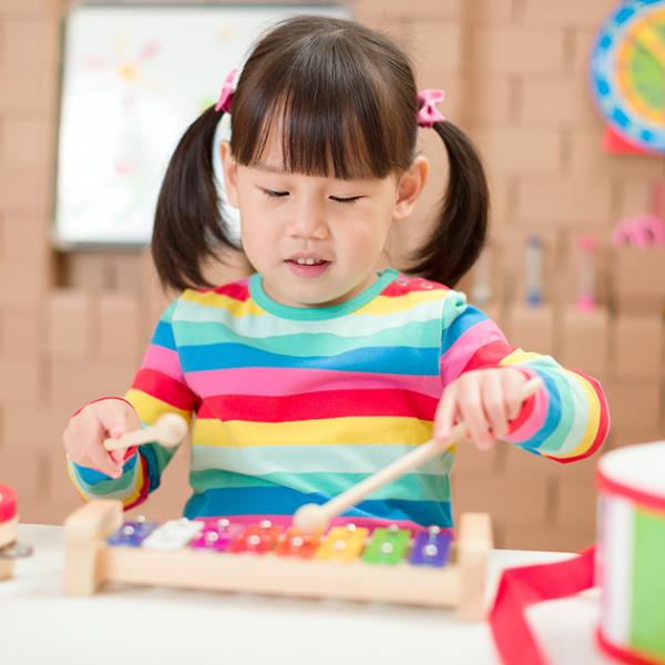 Young girl in a bright striped jumper playing a coloured xylophone, surrounded by musical instruments.