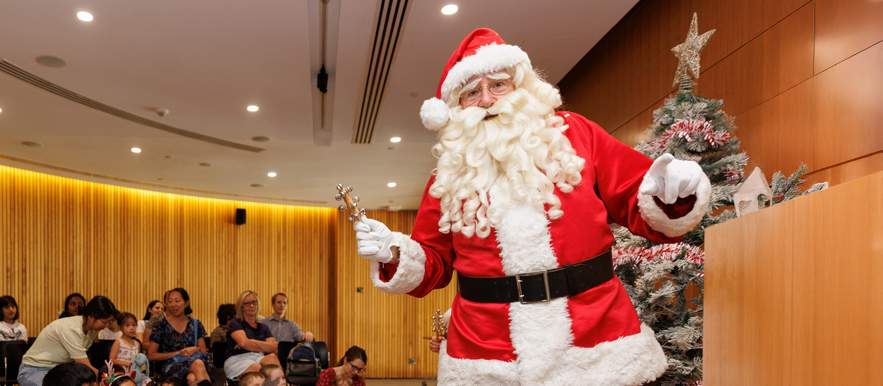 Photo of Santa in the Library auditorium with a crowd of parents and a Christmas tree in the background.