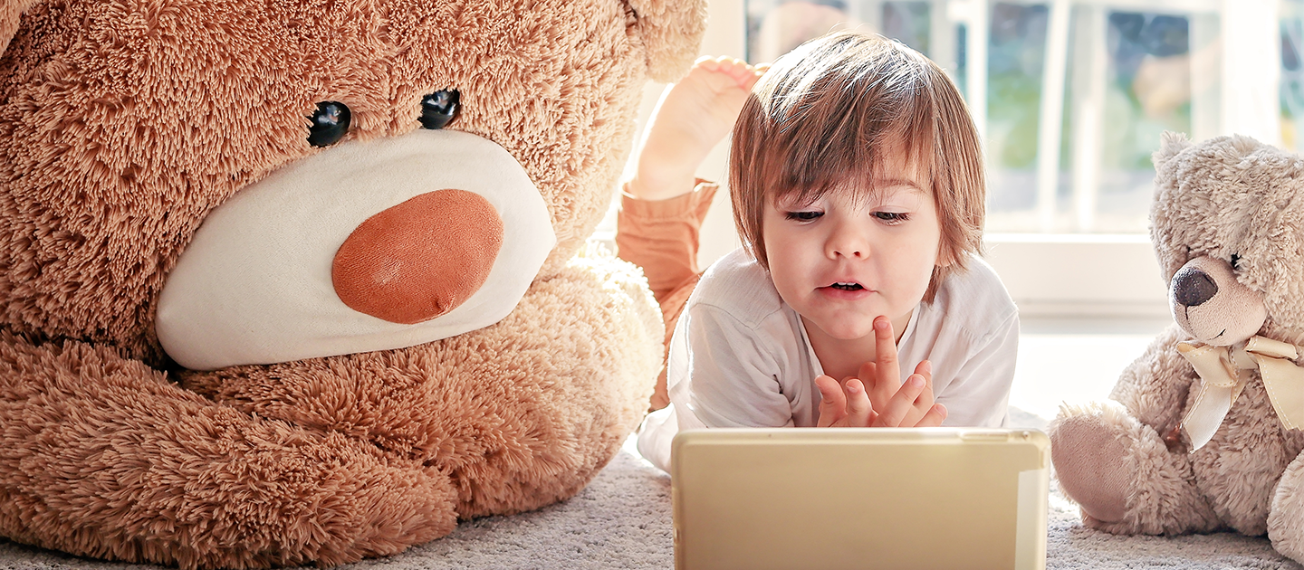 Young child looking at device with a screen between two teddy bears who are also looking at the screen. 