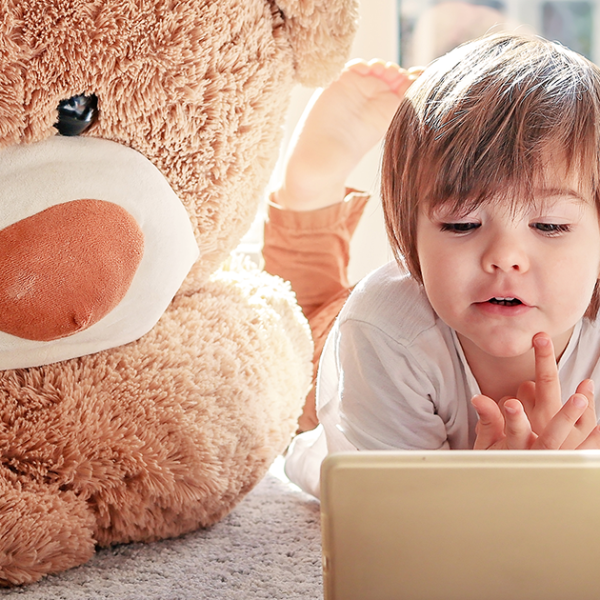 Young child looking at device with a screen between two teddy bears who are also looking at the screen.