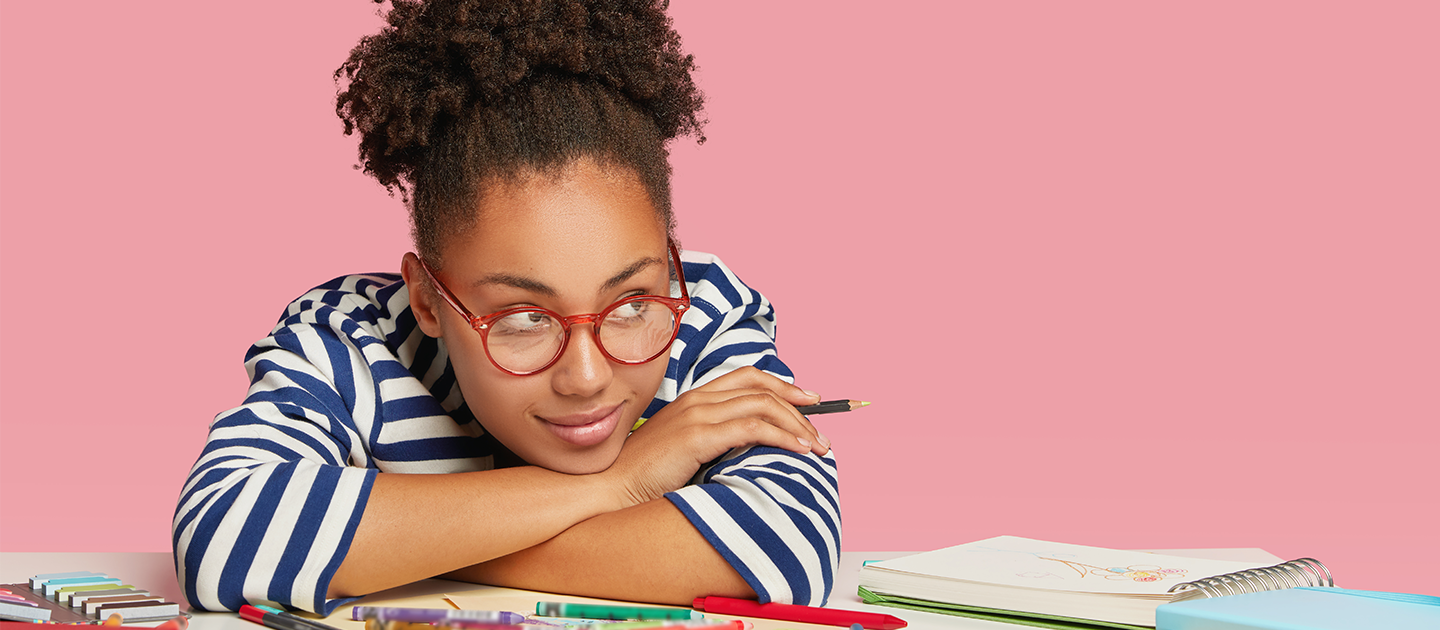 Young adult sitting with her chin on folded arms at a table with stationery and art supplies.