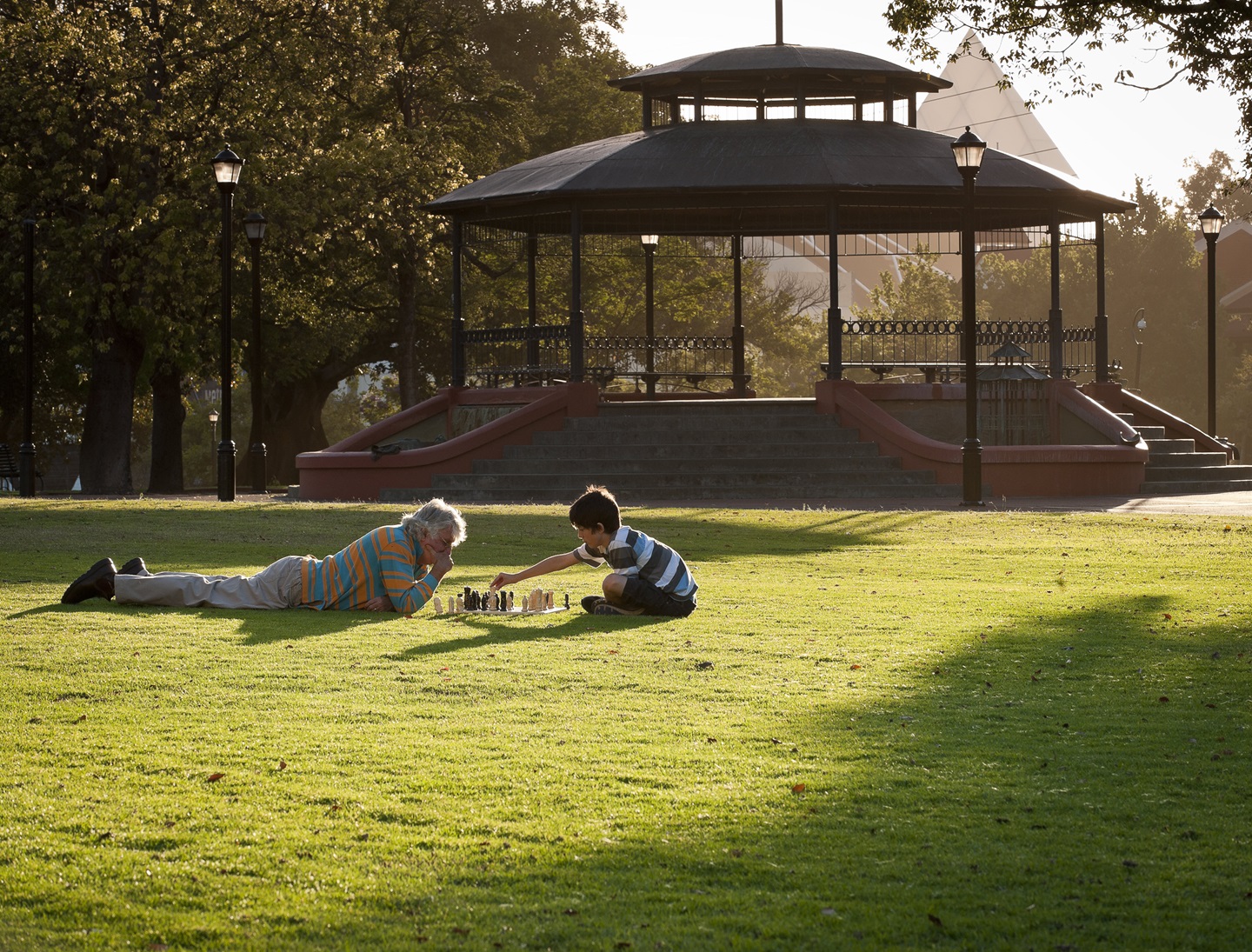 Image of older man and grandchild playing chess on the lawn of Russell Square with the Gazebo in the background